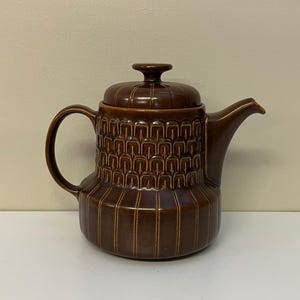 Puede incluir: Una tetera de cerámica marrón con tapa y asa. La tetera tiene un patrón texturizado en el cuerpo y la tapa. El pico es curvo y el asa es un simple lazo. La tetera es probablemente vintage o retro.