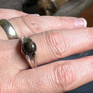 May include: A silver ring with a dark, round gemstone. The ring has a simple, elegant design, with the stone set in a prong setting. Another silver ring is visible on the adjacent finger. The ring is likely a piece of jewelry.