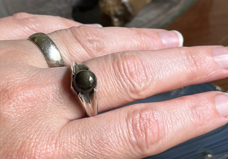 May include: A silver ring with a dark, round gemstone. The ring has a simple, elegant design, with the stone set in a prong setting. Another silver ring is visible on the adjacent finger. The ring is likely a piece of jewelry.