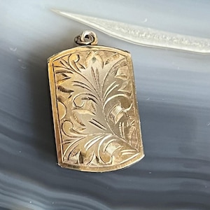 May include: A gold-toned pendant featuring an intricate floral design. The pendant is rectangular in shape and has a smooth, polished finish.