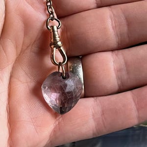 May include: A faceted, heart-shaped pendant in shades of purple and clear, suspended from a gold-colored chain. The pendant is attached to a clasp, and the chain is held in a hand. The jewelry piece is likely a necklace or charm.