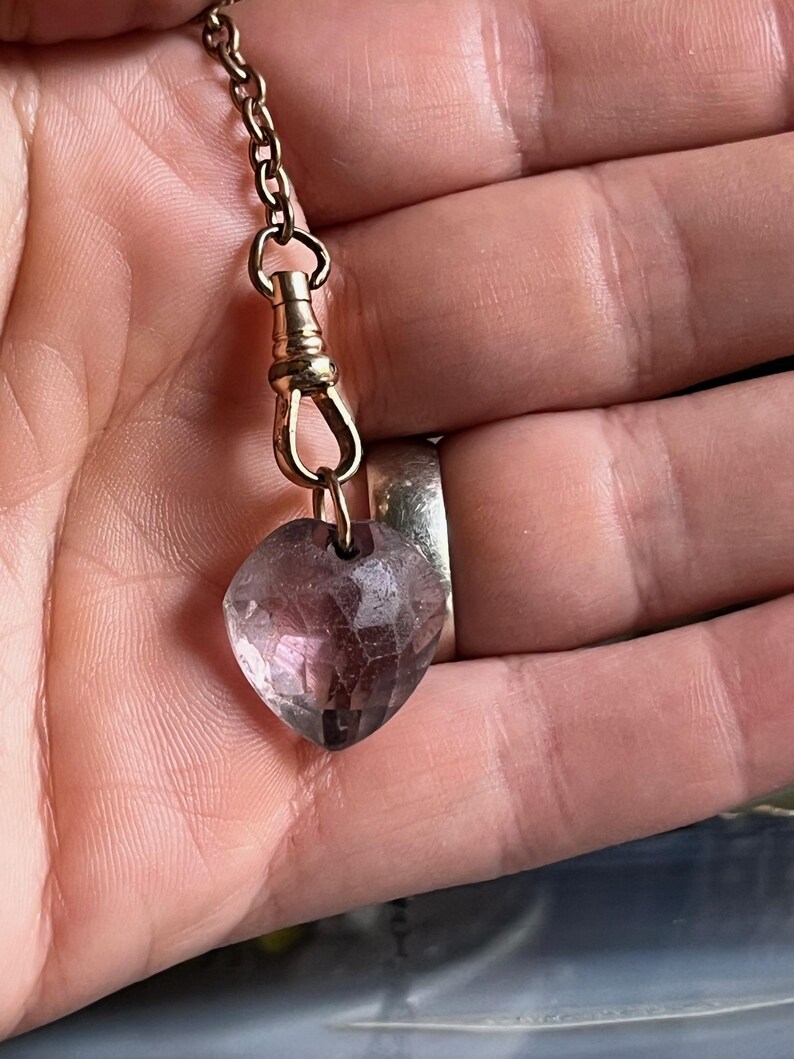 May include: A faceted, heart-shaped pendant in shades of purple and clear, suspended from a gold-colored chain. The pendant is attached to a clasp, and the chain is held in a hand. The jewelry piece is likely a necklace or charm.