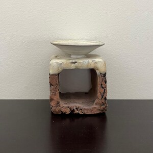 May include: A handcrafted ceramic sculpture featuring a square base with a hollowed-out center. The base is a mix of reddish-brown and beige tones, topped with a small, off-white bowl. The piece has a rustic, textured appearance.