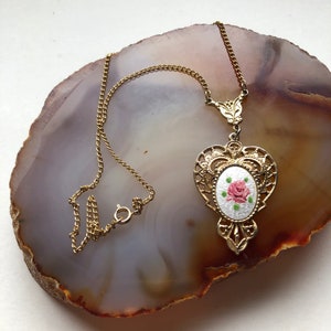 May include: A gold chain necklace with a pendant featuring a white oval with a pink rose design.