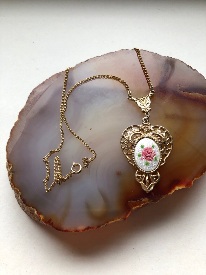 May include: A gold chain necklace with a pendant featuring a white oval with a pink rose design.