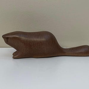 May include: A brown ceramic beaver figurine. The sculpture depicts a beaver in a prone position, with its front paws visible and its tail extended. The figurine has a smooth, glossy finish and is a decorative item.