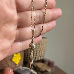 Puede incluir: Un collar de cadena de oro con un colgante de bola texturizada y un borla de cadena colgante.