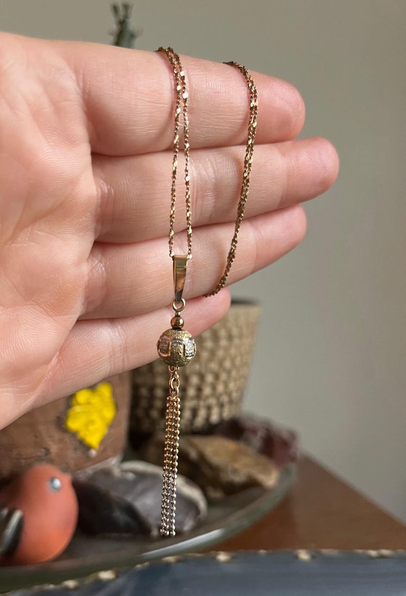 Puede incluir: Un collar de cadena de oro con un colgante de bola texturizada y un borla de cadena colgante.