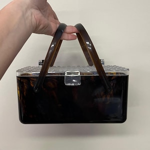 May include: A vintage-style handbag with a tortoiseshell pattern. The rectangular box bag has a clear, textured top and a clear clasp. The handles are a dark brown color. The bag is held by a hand.