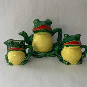 May include: A whimsical ceramic tea set featuring a teapot and two cups, each shaped like a frog. The set is painted with a glossy finish, showcasing a vibrant color palette of green, yellow, and red. The teapot has a lid and a handle.