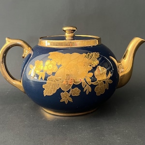 May include: A navy blue teapot with gold detailing. The teapot has a gold handle, spout, and lid. The body is decorated with gold floral patterns. A classic design for tea.
