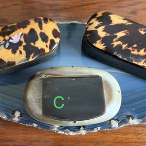 May include: Three vintage tortoiseshell trinket boxes, labeled A, B, and C, are displayed on a blue and white agate slice. Box A features a floral design with a pink flower and silver leaves. Box B is plain and box C is made of horn.