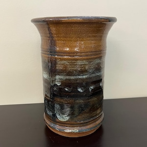 May include: A handmade ceramic vase with a wide opening and a cylindrical body. The vase features a blend of brown, black, and white glaze, creating a textured, layered appearance. The base and rim are a darker brown.