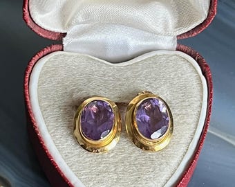 Pode incluir: Um par de brincos de botão dourados com pedras preciosas ovais e roxas. Os brincos são exibidos em uma caixa de joias vermelha em forma de coração com interior branco. A tampa da caixa tem texto dourado.