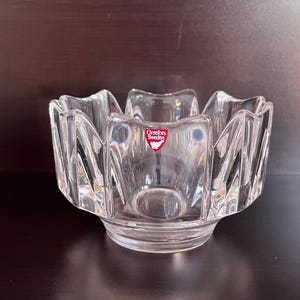 May include: A clear glass bowl with a scalloped edge and a red "Orrefors Sweden" label. The bowl is designed with a unique, faceted pattern.