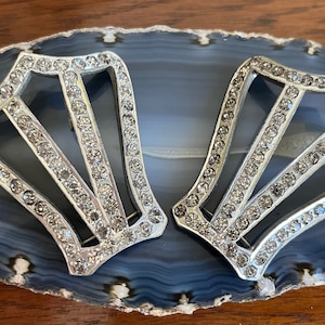 May include: A pair of silver-toned metal shoe buckles with a geometric design and clear rhinestones.