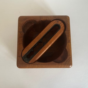 May include: A wooden nutcracker with a metal cracking mechanism, inside a square wooden bowl. The bowl has a carved design and is made of light brown wood.