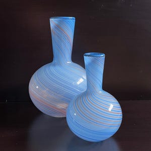 May include: Two glass vases with a light blue and orange striped design. The vases have a round base and a long, slender neck. The larger vase is approximately 10 inches tall, and the smaller vase is about 7 inches tall.