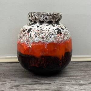 May include: A round, ceramic vase with a textured surface. The vase features a color gradient, transitioning from a dark brown base to a vibrant orange midsection, and a speckled white and black top. The vase has a short neck.
