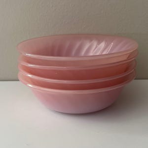 Pode incluir: Uma pilha de quatro tigelas de vidro rosa. As tigelas têm uma forma arredondada com uma borda ligeiramente alargada e um design em espiral no interior. A cor das tigelas é um rosa suave e claro.