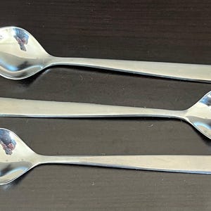 May include: Three stainless steel spoons with rounded bowls and rectangular handles are arranged on a dark surface. The spoons reflect the surrounding environment, showcasing a polished, modern design. These spoons are suitable for serving or eating.