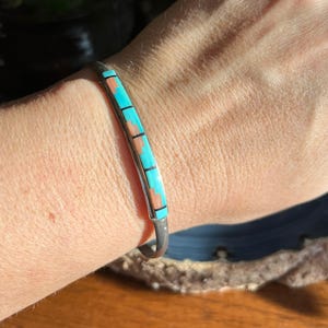 May include: Silver cuff bracelet with turquoise and coral inlay. The bracelet features a rectangular design with alternating turquoise and coral segments, separated by thin black lines. The bracelet is worn on a wrist.