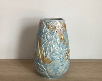 May include: A light blue ceramic vase with a floral design. The vase has a textured surface and is decorated with raised flowers and leaves in shades of brown, yellow, and purple.