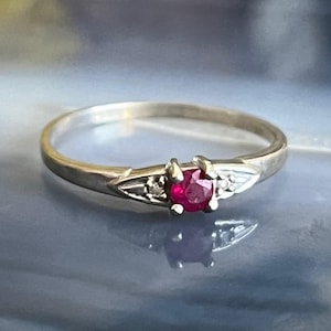 May include: A silver ring featuring a red ruby gemstone with two smaller white gemstones flanking it.