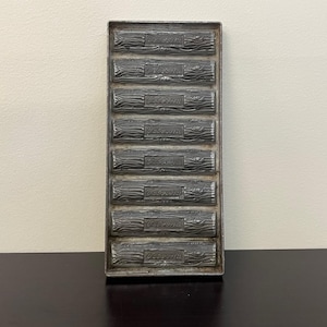 May include: A rectangular metal mould with seven log-shaped compartments. Each log has the word "Welcome" embossed on it. The mould has a rustic, antique appearance with a dark, aged finish, suggesting it is vintage.
