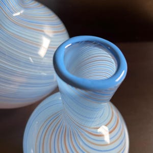 May include: Two glass vases with a light blue base and a pattern of thin, swirling lines in orange and blue. The rim of the vase is a solid light blue. The vases have a bulbous body and a narrow neck.