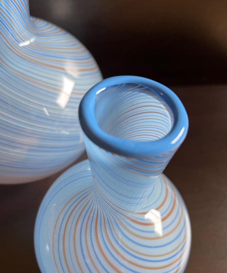 May include: Two glass vases with a light blue base and a pattern of thin, swirling lines in orange and blue. The rim of the vase is a solid light blue. The vases have a bulbous body and a narrow neck.