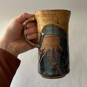 Può includere: Una tazza alta in ceramica fatta a mano con un manico grande. La tazza presenta un design strutturato e terroso con una miscela di colori marrone, nero e beige. La parte superiore è beige, che passa a un motivo scuro e astratto nella metà inferiore.