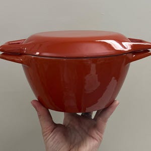 May include: A bright orange Dutch oven with a matching lid. The pot has two side handles and a rounded shape. The glossy finish reflects light, highlighting the cookware's smooth surface. This kitchenware is ideal for cooking.