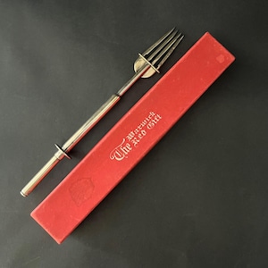 Puede incluir: Un tenedor de servicio de metal plateado con un mango largo y delgado y un protector circular, colocado junto a una caja de presentación rectangular roja. La caja presenta el texto en color dorado "The Warwick The Red Gift" en la tapa.