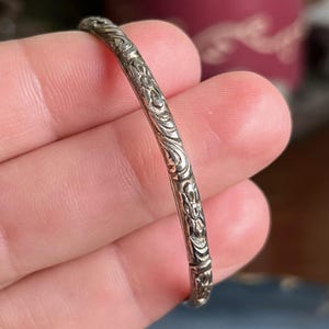Peut inclure: Un bracelet jonc argenté avec un motif floral et volute complexe. Le bracelet est fin et semble être en métal. Les détails du motif sont visibles, mettant en valeur le savoir-faire de la pièce.