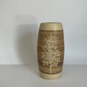 May include: A ceramic vase with a cylindrical shape, featuring a textured brown and beige design. The vase has a detailed tree motif carved into the front. The top and bottom rims are a lighter beige colour.