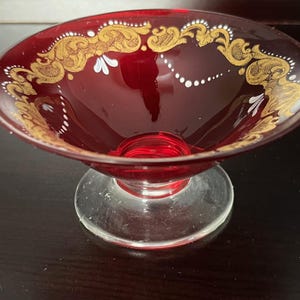 Puede incluir: Un cuenco de cristal rojo rubí con una base clara y circular. El borde está decorado con filigranas doradas y detalles blancos. Este cuenco decorativo es ideal para servir fruta o como pieza decorativa.
