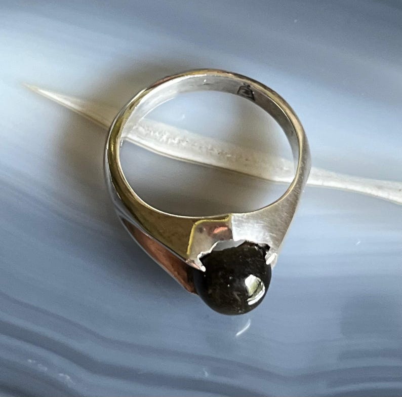 May include: A silver ring with a dark, rounded gemstone. The ring has a unique design with a geometric shape on the band. The gemstone is set in a bezel setting, and the ring is displayed on a light blue surface.