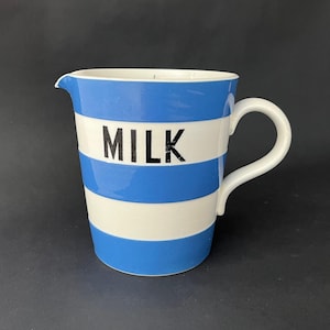 May include: A ceramic milk pitcher with a blue and white striped design. The word "MILK" is printed in black block letters on the front. The pitcher has a handle and a spout for easy pouring.