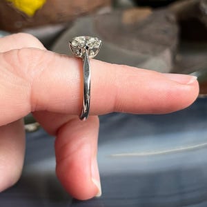 Puede incluir: Un anillo de plata con una piedra preciosa redonda y transparente engastada en garras. El anillo est&aacute; en un dedo.