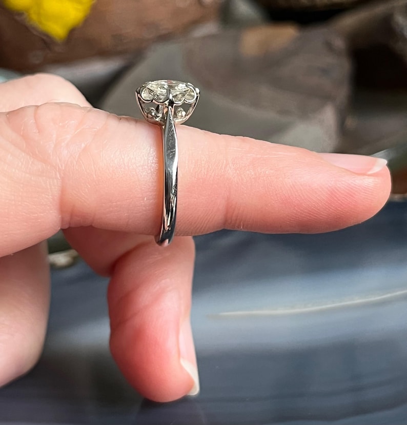 Puede incluir: Un anillo de plata con una piedra preciosa redonda y transparente engastada en garras. El anillo est&aacute; en un dedo.