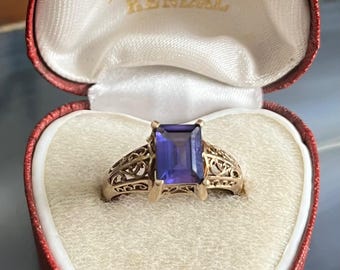 May include: A gold ring with a filigree design and a large, rectangular, purple gemstone. The ring is in a red heart-shaped box with the words "H. Rogantin" and "Kendal" printed on the inside.