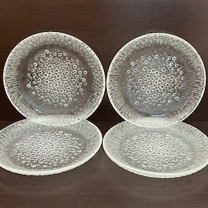 May include: Four clear glass plates with a textured, bubble-like pattern. Two plates are stacked on top of the other two. The plates are round with a slightly raised edge, suitable for serving food.
