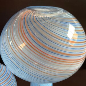 May include: A spherical glass object with a light blue base and a swirling pattern of orange, blue, and white stripes. The glass has a glossy finish and reflects light, creating a visually appealing decorative piece.