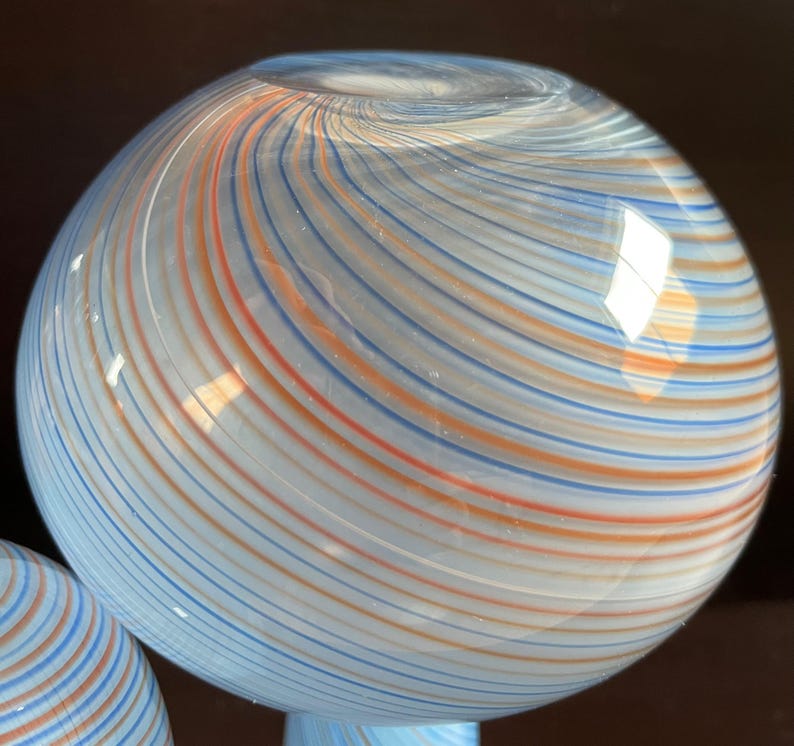 May include: A spherical glass object with a light blue base and a swirling pattern of orange, blue, and white stripes. The glass has a glossy finish and reflects light, creating a visually appealing decorative piece.