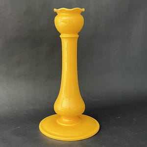 May include: A tall, yellow glass candlestick holder with a round base, a slender stem, and a flared top. The candlestick is a vibrant shade of yellow and has a glossy finish. The design is simple and elegant, suitable for various decor styles.