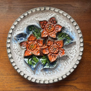 May include: A round, ceramic wall hanging featuring a raised floral design. The flowers are orange with green leaves and blue accents, set against a textured white background. The border is a series of small, raised dots.