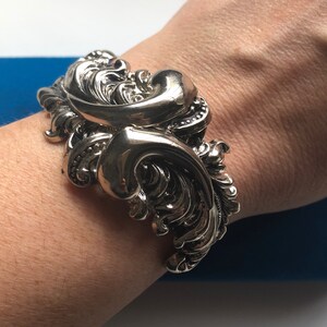 K&ouml;nnte beinhalten: Ein silberfarbenes Armreif mit einem verzierten, wirbelnden Design. Das Armband ist breit und hat eine strukturierte Oberfl&auml;che.