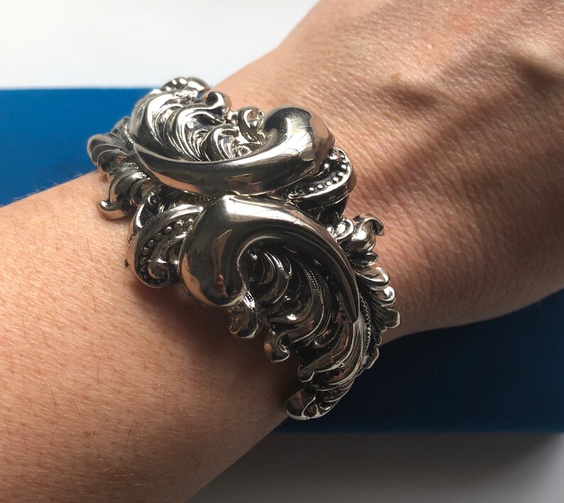 K&ouml;nnte beinhalten: Ein silberfarbenes Armreif mit einem verzierten, wirbelnden Design. Das Armband ist breit und hat eine strukturierte Oberfl&auml;che.