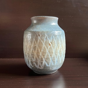 May include: A handmade ceramic vase with a diamond pattern carved into the surface. The vase has a light blue and tan colour scheme, with a slightly textured finish. The vase has a rounded body and a narrow neck, suitable for displaying flowers.